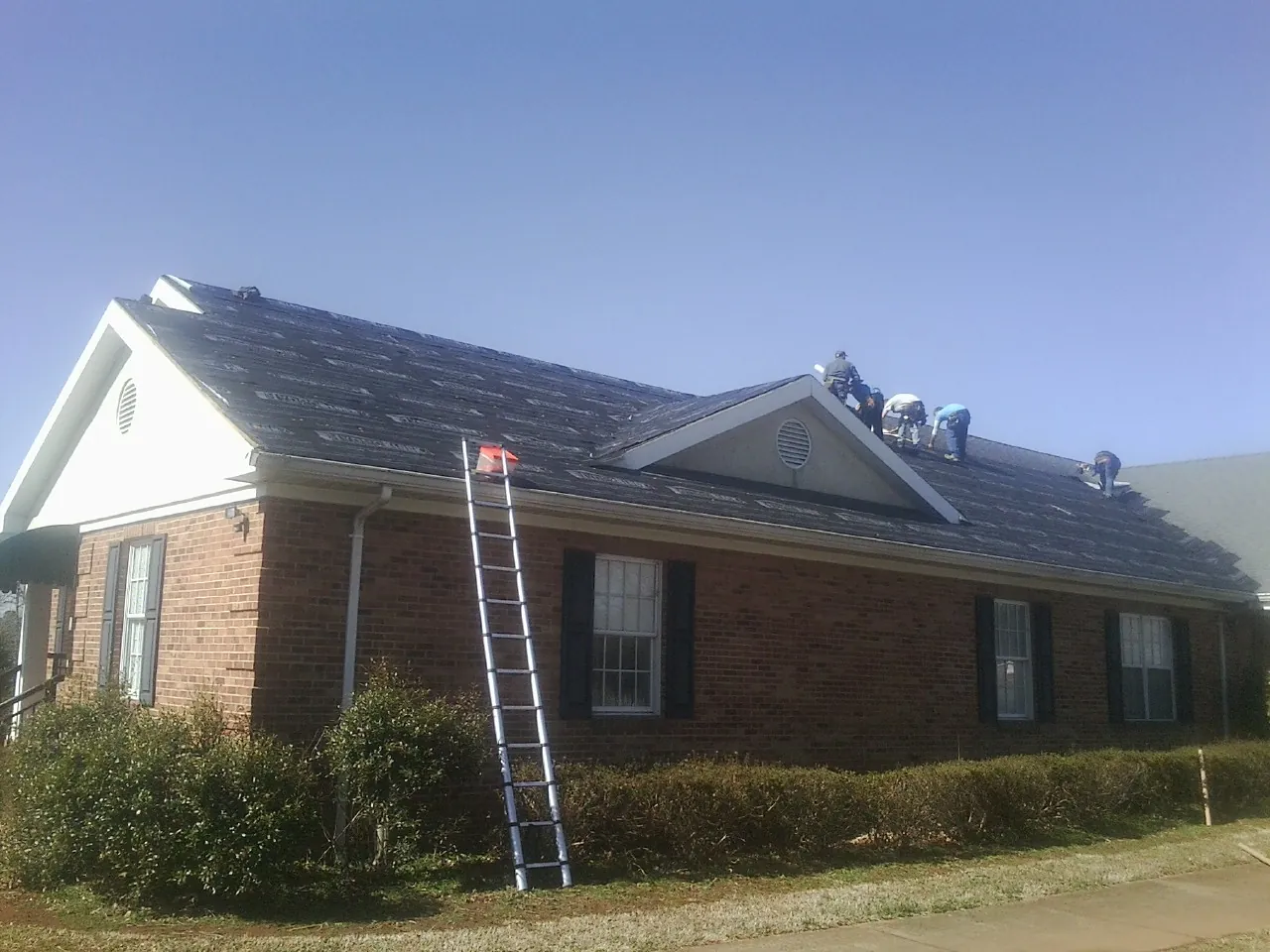 Crew tearing shingles off a brick ranch roof with a ladder set — Emergency Roof Repair work by our crew in Basco