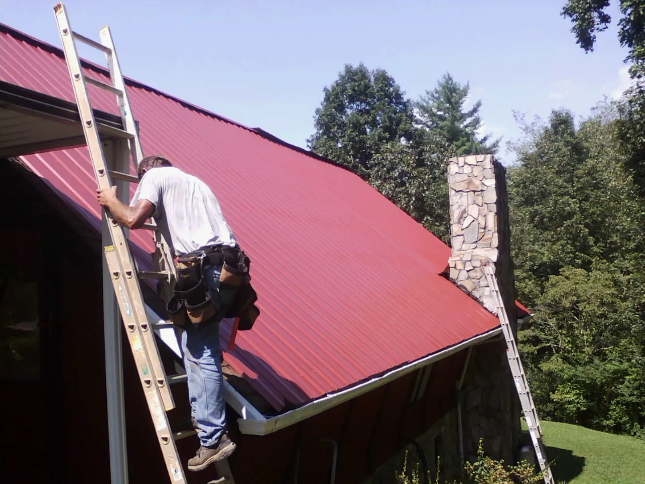Roofer on a ladder beside a red metal roof and stone chimney — Soffit & Fascia Repair by Roofing Brothers in Basco